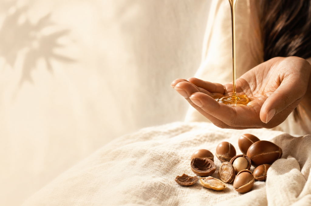 Warm golden argan oil pouring into a cupped palm beside argan kernels on ivory linen