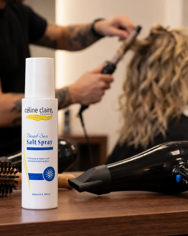 Celine Claire Dead Sea Salt Spray 200 ml bottle on a salon station beside a round brush and blow-dryer, with hair being styled in the background